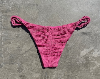Puede incluir: Braguita de bikini magenta con tejido texturizado y fruncido. Los laterales presentan gruesas correas trenzadas. La prenda está diseñada con una pernera de corte alto y una cobertura trasera moderada, adecuada para trajes de baño.