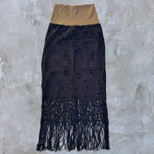 May include: A black skirt with a gold waistband and fringe trim. The skirt features a detailed, openwork design. The fringe hangs from the bottom hem. The skirt is displayed on a neutral background.