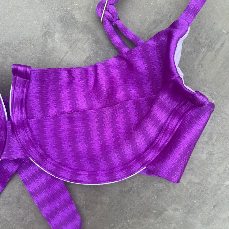 May include: Purple satin bikini top with a striped pattern. The top has a halter style with adjustable straps and a clasp closure.