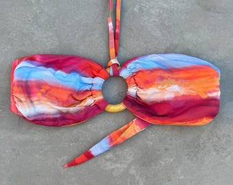 May include: A colorful bandeau bikini top with a sunset-inspired design. The top features shades of red, orange, and blue, with a central gray stone detail and a yellow ring. The straps are multi-colored.