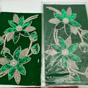 May include: Two green fabric flower appliques with silver sequins and beads on a green felt background. The flowers have five petals each and are arranged in a floral pattern.