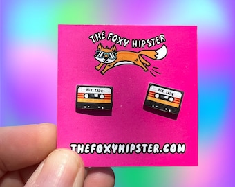 May include: A pair of cassette tape earrings on a pink card with the text "The Foxy Hipster" and "thefoxyhipster.com". The earrings are black with a yellow and orange cassette tape design and the text "Mix Tape" on them.