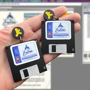 May include: A pair of black and silver floppy disk earrings with a blue and white America Online logo and a yellow stick figure. The earrings are hanging from a person's hand.