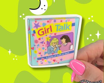 May include: A sticker featuring the board game "Girl Talk." The sticker has a pink, blue, and yellow color scheme. The game's cover art shows two girls talking. The text "Girl Talk" is in yellow. The sticker is held by a hand with pink nail polish.