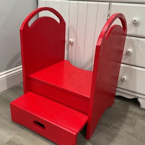 May include: A bright red wooden step stool with two steps and a handle on each side. The step stool is designed to help children reach higher surfaces. The stool has a simple, functional design.
