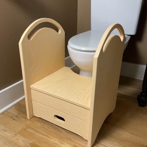 May include: A light-colored wooden step stool featuring two tall side panels with integrated arched handles and a lower drawer with a horizontal oval cutout. The stool has a flat top surface and is constructed from light-toned wood with a visible grain.