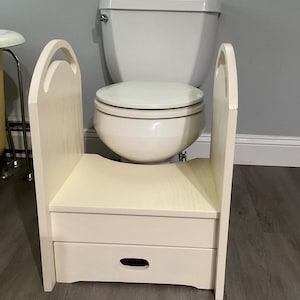 May include: A cream-colored wooden step stool with a drawer, designed for use in a bathroom. The stool has a rectangular seat and two arched handles. It is positioned in front of a white toilet.