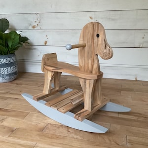 May include: A wooden rocking horse with a light brown finish. The horse has a gray base and a small, round, gray handle. The horse is made of wood and has a simple design, suitable for young children. The horse is a classic toy.