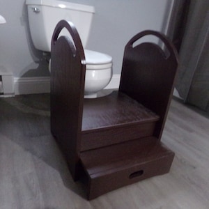 May include: A dark brown wooden step stool with two steps and a handle on each side. The step stool is designed to help children reach the sink or toilet. The stool is in a bathroom setting.