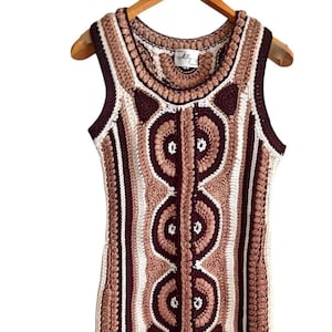 May include: A sleeveless, crochet dress in shades of cream, brown, and burgundy. The dress features a repeating pattern of large, stylized circles and vertical stripes. The dress is hanging on a wooden hanger.