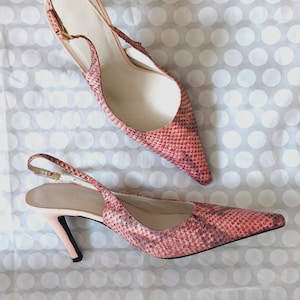Pode incluir: Um par de sapatos de salto rosa com padrão de pele de cobra e bico fino. Os sapatos têm uma fina tira no tornozelo com fivela dourada e um salto pequeno. Os sapatos estão sobre um fundo branco com bolinhas.