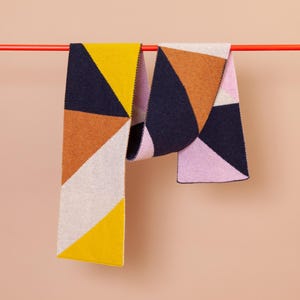 Könnte beinhalten: Zwei geometrisch gemusterte Schals hängen an einer roten Stange. Die Schals sind aus weichem, gestricktem Stoff und haben eine Mischung aus gelben, orangefarbenen, rosa, weißen und dunkelblauen Farben.
