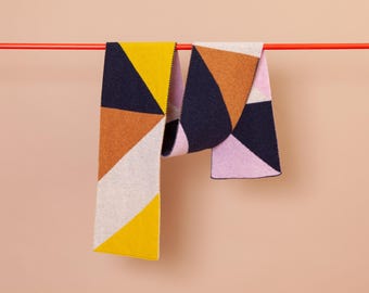 May include: Two geometric patterned scarves hanging on a red rod. The scarves are made of a soft, knitted fabric and feature a mix of yellow, orange, pink, white, and navy blue colors.