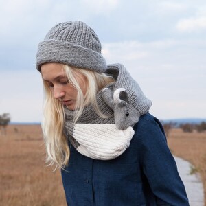 May include: A woman wearing a gray knitted hat and a gray and white knitted scarf with a small gray animal attached. The scarf is worn around the neck and the animal is hanging down the front of the scarf.