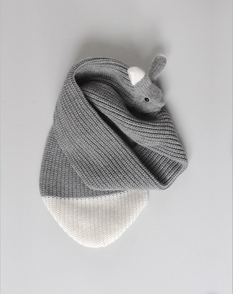 May include: Gray and white knitted scarf with a gray rabbit head on one end. The rabbit has white ears and black eyes.