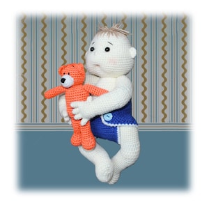 Op de afbeelding: Een gehaakte pop met blond haar die een blauwe luier draagt en een gehaakte oranje teddybeer vasthoudt.