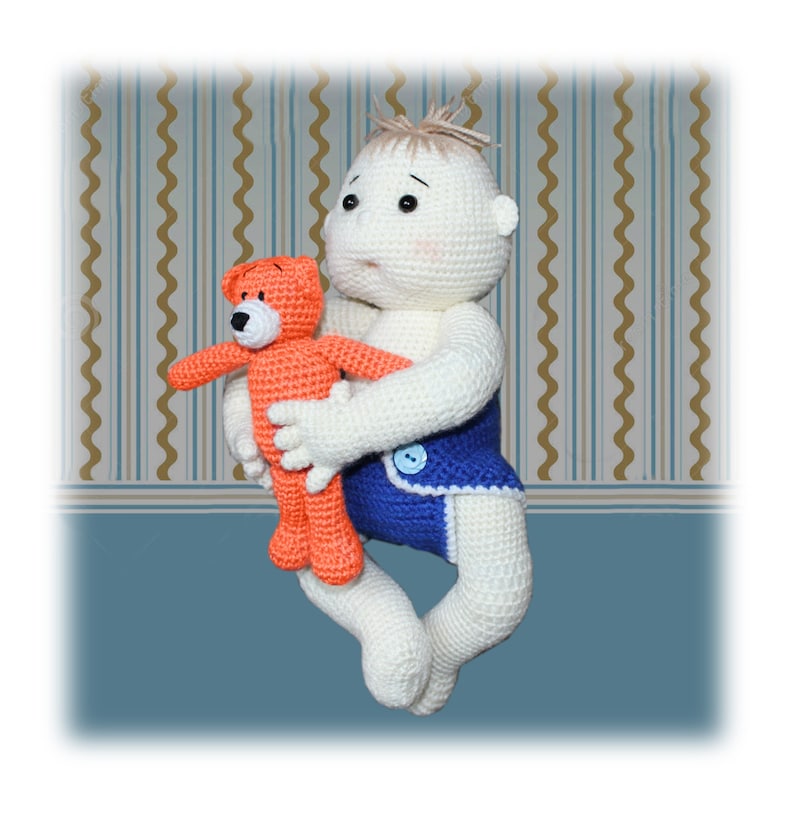 Op de afbeelding: Een gehaakte pop met blond haar die een blauwe luier draagt en een gehaakte oranje teddybeer vasthoudt.