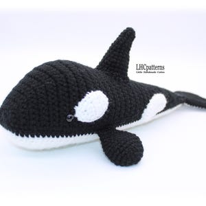 May include: A handcrafted crochet orca whale in black and white. The soft toy has a black body with white markings, a dorsal fin, and a tail. The text "LHCpatterns" is visible on the side of the whale.