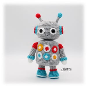 May include: A handmade grey crochet robot toy with red, blue, white, and yellow accents. The robot has large black eyes, antennae, and colourful details on its body and limbs. The text "LHCpatterns" is visible.