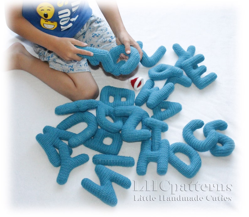 May include: A set of blue crocheted alphabet letters. The letters are arranged on a white surface. The letters are soft and cuddly, perfect for a child's playroom.