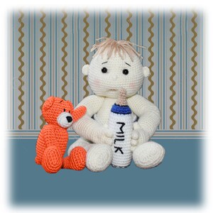 Op de afbeelding: Een witte gehaakte babypop met blond haar zit op een blauw en bruin gepatroneerd oppervlak. De pop houdt een blauwe en witte fles vast waarop "MILK" staat. Een kleine oranje gehaakte teddybeer zit naast de pop.
