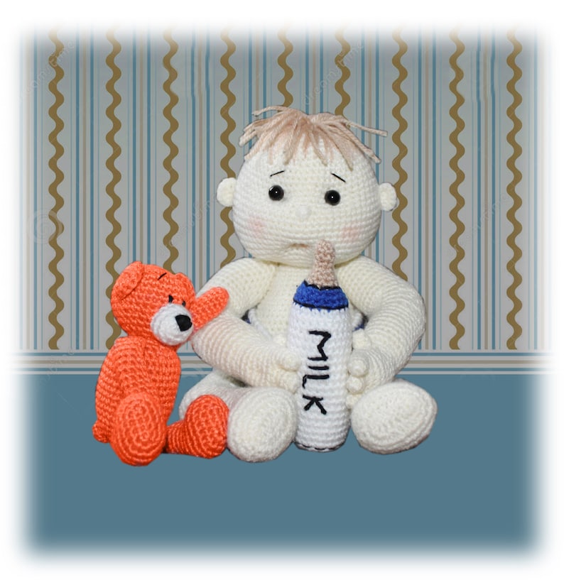 Op de afbeelding: Een witte gehaakte babypop met blond haar zit op een blauw en bruin gepatroneerd oppervlak. De pop houdt een blauwe en witte fles vast waarop "MILK" staat. Een kleine oranje gehaakte teddybeer zit naast de pop.