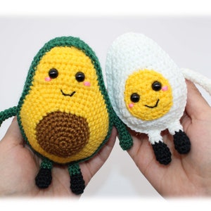 May include: Two crocheted plush toys, one shaped like an avocado and the other shaped like an egg. The avocado is green, yellow, and brown, and the egg is white and yellow. Both toys have smiling faces with black eyes and pink cheeks.