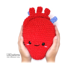 May include: A red crocheted heart-shaped plush toy with a smiling face and blue crocheted details. The toy is being held by two hands.