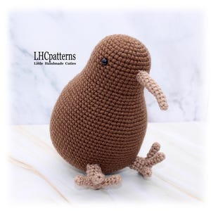 May include: A handmade brown crochet kiwi bird with a long beak and small feet. The bird has a round body and a small black eye. The text "LHCpatterns Little Handmade Cuties" is in the upper left corner.