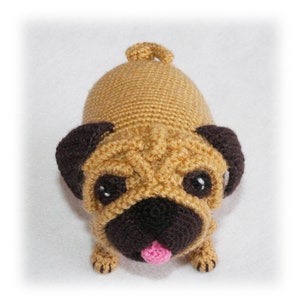 May include: A handmade crochet pug dog toy. The pug is tan with dark brown ears and muzzle. It has black eyes and a pink tongue. The toy is made with a textured stitch and has a small tail.