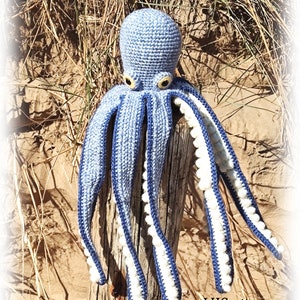 May include: A crocheted blue octopus with white pom-poms on the tentacles. The octopus is sitting on a wooden post in a sandy area with grass.