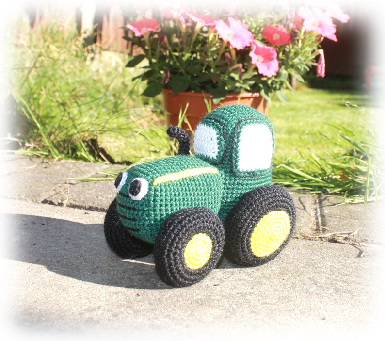 May include: A green crocheted toy tractor with black wheels and yellow accents. The tractor has a white windscreen and black details.