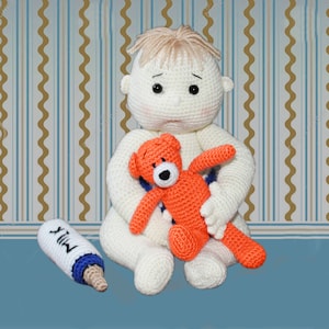 Op de afbeelding: Een gehaakte babypop met blond haar, gekleed in een witte outfit, houdt een gehaakte oranje teddybeer vast. Een klein wit flesje met het woord "MILK" erop staat op het blauwe oppervlak.
