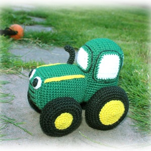 May include: A green crocheted toy tractor with black wheels and yellow tyre treads. The tractor has a white crocheted windscreen and black crocheted details.