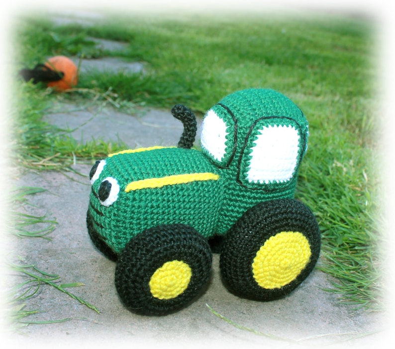 May include: A green crocheted toy tractor with black wheels and yellow tyre treads. The tractor has a white crocheted windscreen and black crocheted details.