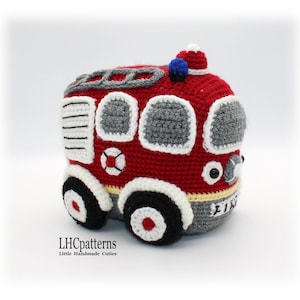 May include: A red, gray, and white crocheted fire truck toy with black wheels and a blue light on top. The truck has a white stripe around the bottom and the word "FIRE" written on the side.