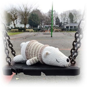 Op de afbeelding: Een wit gebreid pluche speelgoed in de vorm van een beer met een bruin en wit gestreepte trui, liggend op een zwarte schommel.