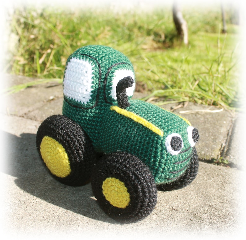 May include: A green crocheted toy tractor with black wheels and yellow accents. The tractor has white eyes and a white window.
