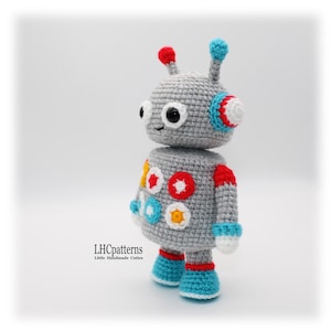 May include: A handmade gray crochet robot toy with red, blue, white, and yellow accents. The robot has a round head with antennae, large eyes, and colorful circular details on its body. The text "LHCpatterns" is visible.