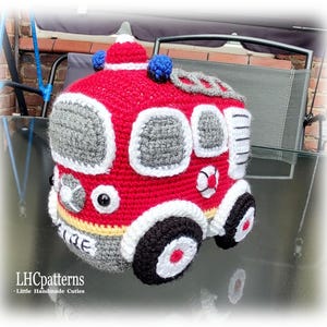 May include: A red, white and grey crocheted fire engine toy with black wheels. The toy has a yellow stripe and a blue light on top.
