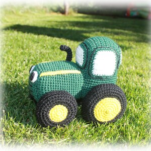May include: A green and black crocheted toy tractor with yellow wheels. The tractor has a white window and black details.
