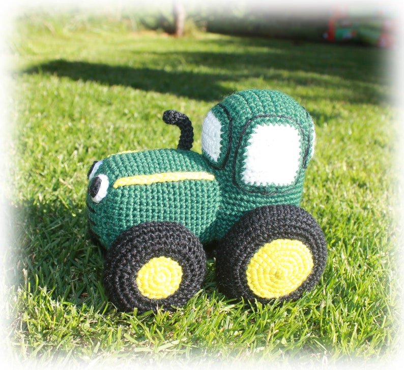 May include: A green and black crocheted toy tractor with yellow wheels. The tractor has a white window and black details.
