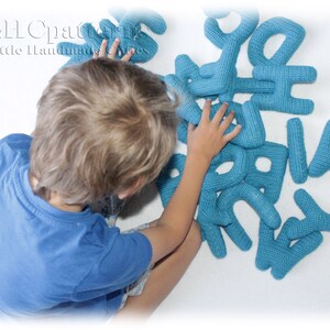 May include: A child plays with a set of blue crocheted alphabet letters. The letters are made of yarn and are soft and cuddly. The letters are arranged in a random order on a white surface.