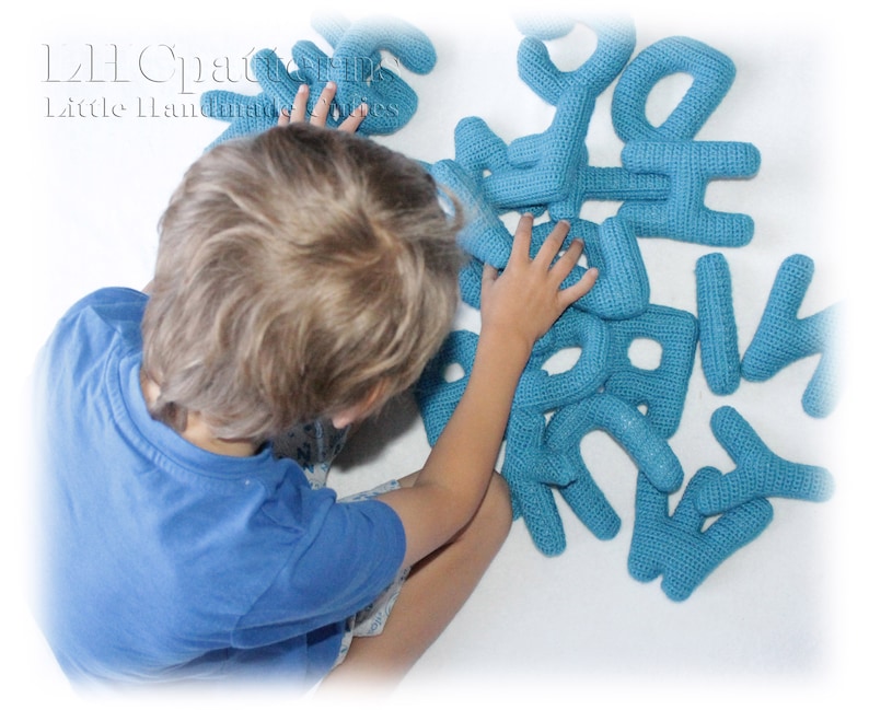 May include: A child plays with a set of blue crocheted alphabet letters. The letters are made of yarn and are soft and cuddly. The letters are arranged in a random order on a white surface.