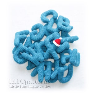 May include: A collection of blue crocheted letters, some with a white outline, are scattered on a white background. A small red crocheted Santa hat is placed on top of the letters. The letters spell out "L2L Patterns Little Handmade Cuties".