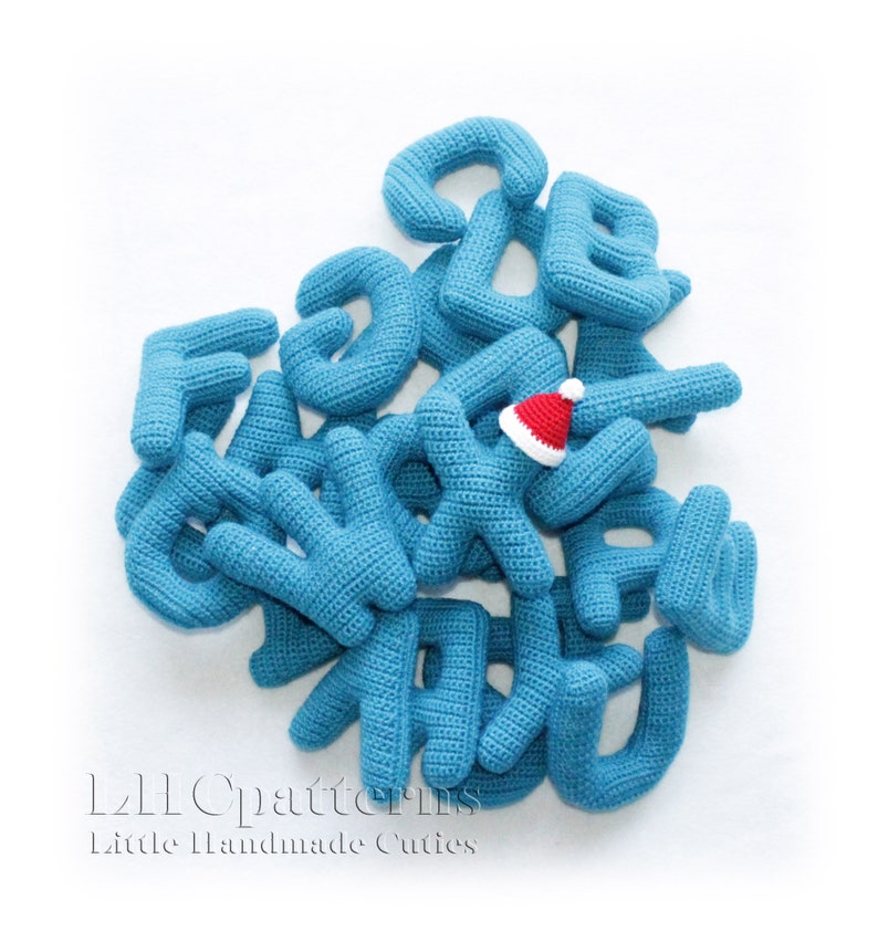 May include: A collection of blue crocheted letters, some with a white outline, are scattered on a white background. A small red crocheted Santa hat is placed on top of the letters. The letters spell out "L2L Patterns Little Handmade Cuties".