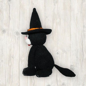 May include: A black crocheted cat wearing a witch's hat with an orange band. The cat is sitting on a white wooden surface.