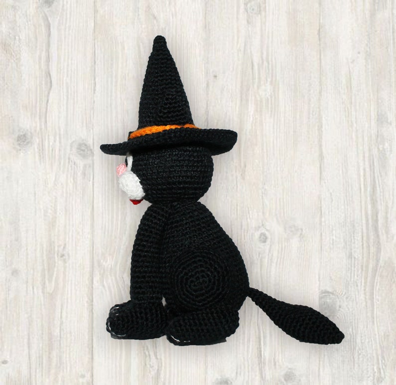 May include: A black crocheted cat wearing a witch's hat with an orange band. The cat is sitting on a white wooden surface.