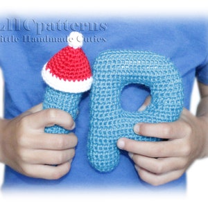 May include: A blue crocheted letter P with a red and white crocheted Santa hat on top. The letter is being held by a person's hands.