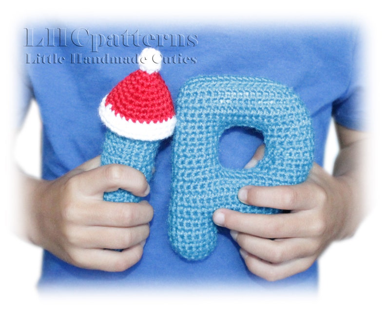 May include: A blue crocheted letter P with a red and white crocheted Santa hat on top. The letter is being held by a person's hands.
